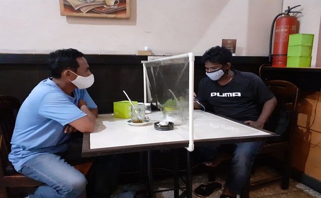 Dua pengunjung Rumah Kopi Lela, duduk dibatasi alat pembatas. Mereka juga menggunakan masker. Foto: Beritabeta